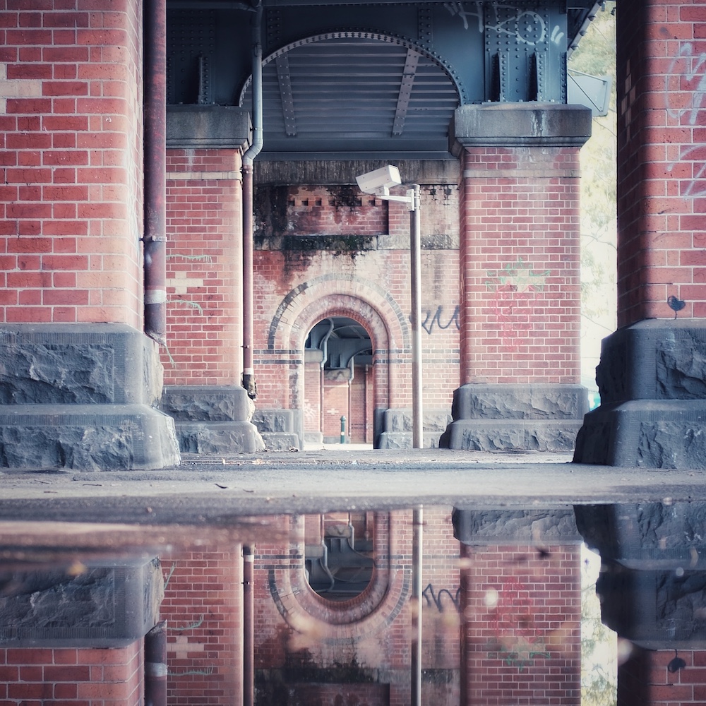 Some old brick arches under an overpass. The foreground is a puddle reflecting the background.
