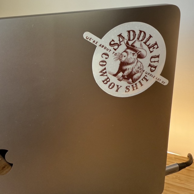 A Macbook with a sticker on it. The sticker is a Capybara in a cowboy hat and the lettering says ‘Saddle up, we’re about to do a whole lot of cowboy shit